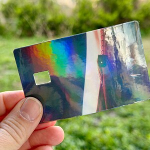 Puede incluir: Una tarjeta rectangular iridiscente con un recorte cuadrado, sostenida en una mano. La superficie de la tarjeta refleja un espectro de colores, incluyendo azules, verdes y amarillos. El fondo est&aacute; borroso, con follaje verde.