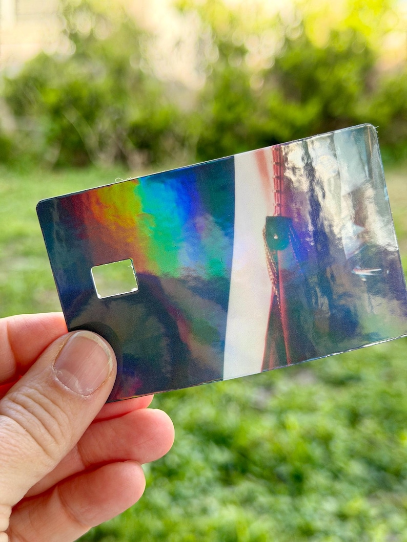 Puede incluir: Una tarjeta rectangular iridiscente con un recorte cuadrado, sostenida en una mano. La superficie de la tarjeta refleja un espectro de colores, incluyendo azules, verdes y amarillos. El fondo est&aacute; borroso, con follaje verde.