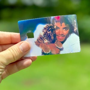 Peut inclure: Une carte rectangulaire en plastique pr&eacute;sentant une image holographique d'une personne aux cheveux boucl&eacute;s fonc&eacute;s tenant un petit lionceau. La carte comporte une d&eacute;coupe carr&eacute;e sur le c&ocirc;t&eacute; gauche et affiche un reflet lumineux multicolore.