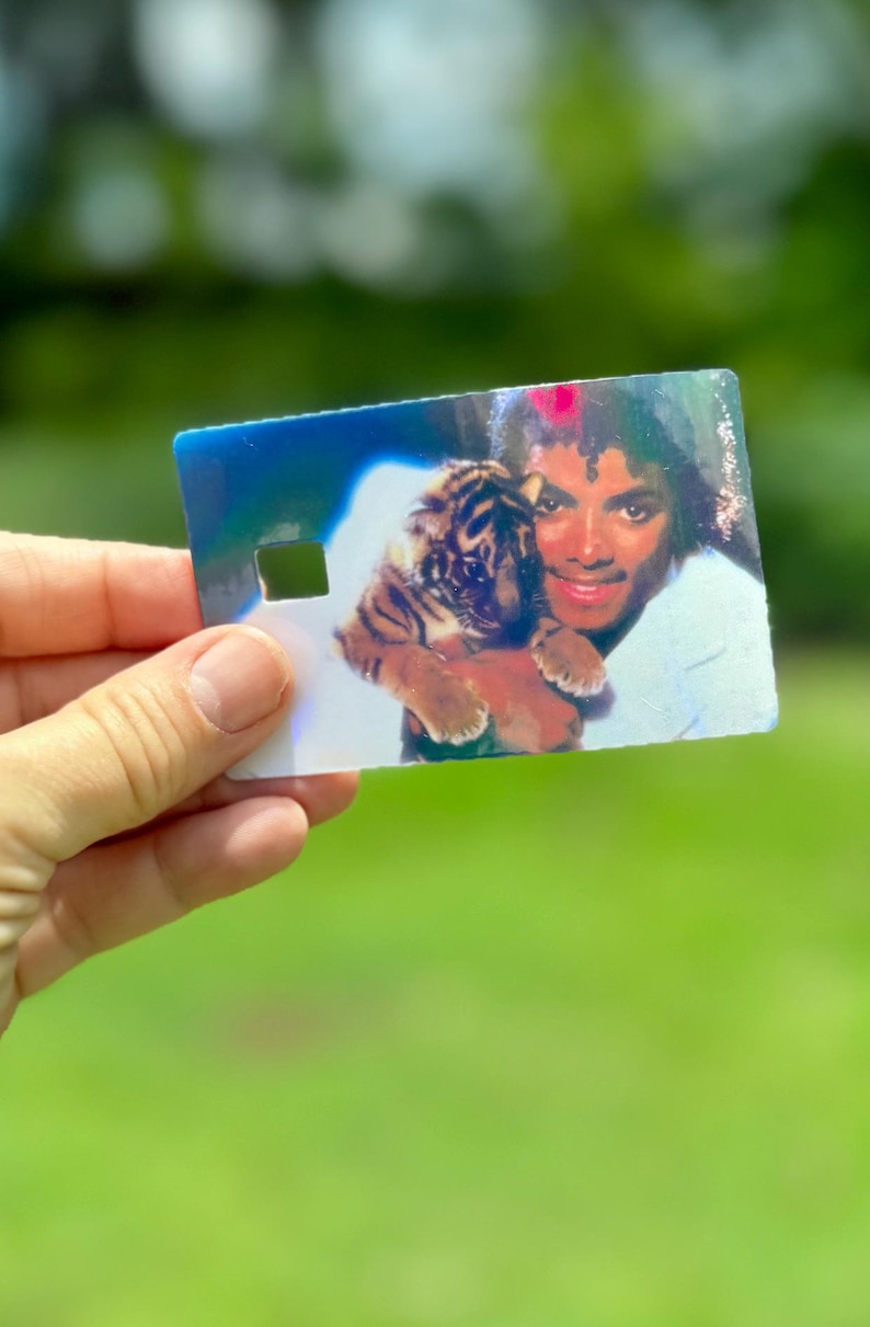 Peut inclure: Une carte rectangulaire en plastique pr&eacute;sentant une image holographique d'une personne aux cheveux boucl&eacute;s fonc&eacute;s tenant un petit lionceau. La carte comporte une d&eacute;coupe carr&eacute;e sur le c&ocirc;t&eacute; gauche et affiche un reflet lumineux multicolore.