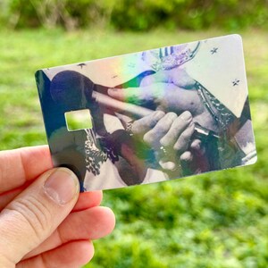 Puede incluir: Un art&iacute;culo hologr&aacute;fico del tama&ntilde;o de una tarjeta de cr&eacute;dito con una imagen en blanco y negro de manos sosteniendo un micr&oacute;fono. La tarjeta tiene un recorte rectangular y un brillo reflectante, similar al arco&iacute;ris.