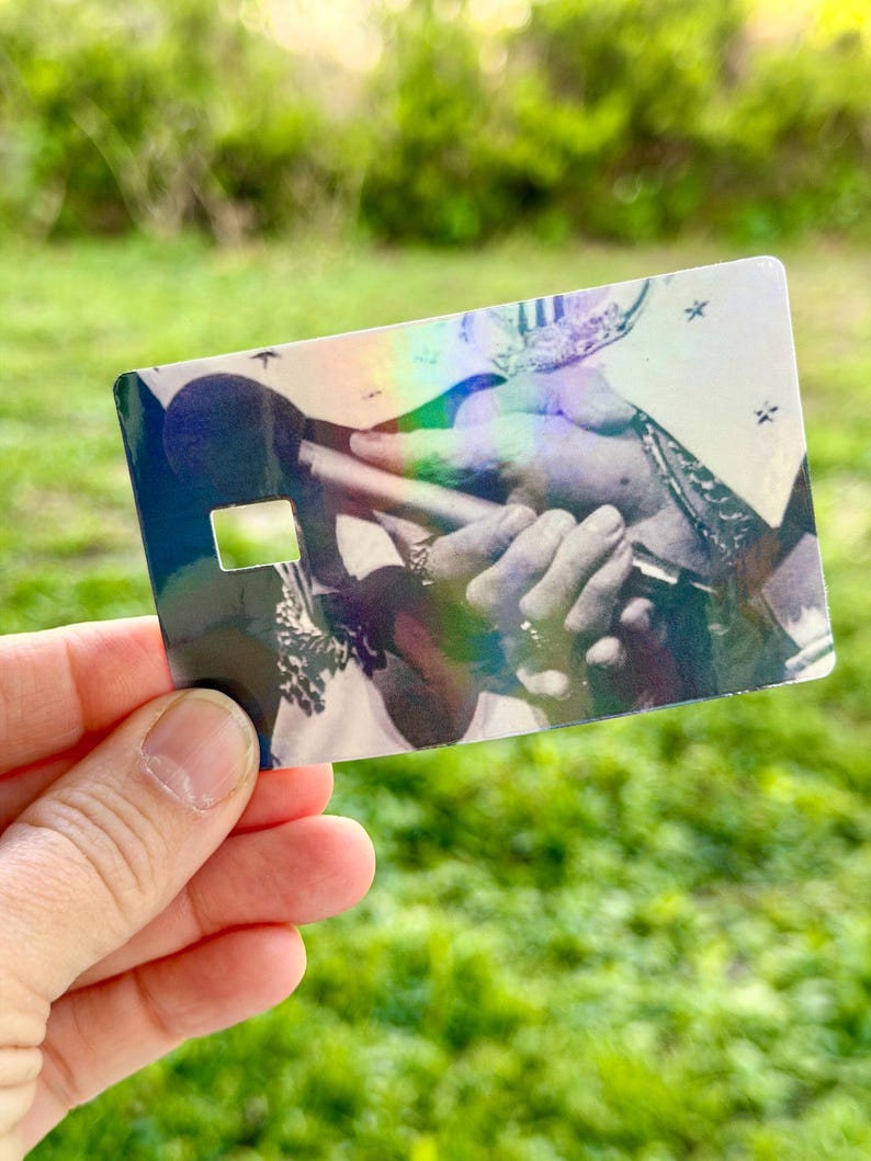 Puede incluir: Un art&iacute;culo hologr&aacute;fico del tama&ntilde;o de una tarjeta de cr&eacute;dito con una imagen en blanco y negro de manos sosteniendo un micr&oacute;fono. La tarjeta tiene un recorte rectangular y un brillo reflectante, similar al arco&iacute;ris.