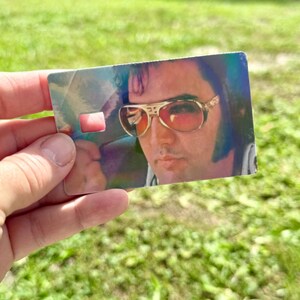 Puede incluir: Una tarjeta rectangular con un retrato de Elvis Presley con gafas de sol con montura dorada y lentes rojas. La tarjeta tiene un brillo hologr&aacute;fico y un peque&ntilde;o recorte rectangular. La imagen se sostiene contra un fondo verde borroso.