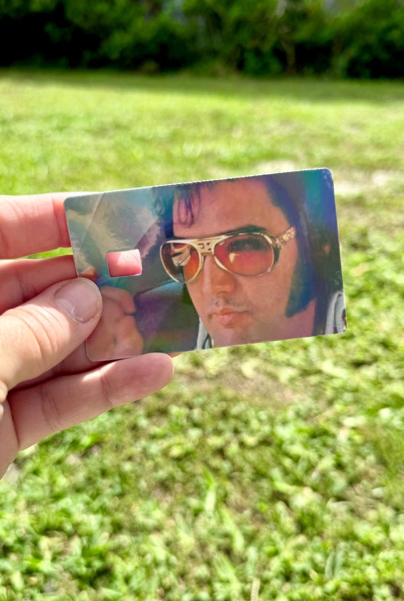 Puede incluir: Una tarjeta rectangular con un retrato de Elvis Presley con gafas de sol con montura dorada y lentes rojas. La tarjeta tiene un brillo hologr&aacute;fico y un peque&ntilde;o recorte rectangular. La imagen se sostiene contra un fondo verde borroso.