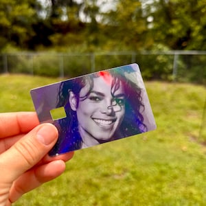 Peut inclure: Une carte rectangulaire en plastique pr&eacute;sentant un portrait photographique en noir et blanc de Michael Jackson. La carte poss&egrave;de une finition holographique qui refl&egrave;te la lumi&egrave;re, cr&eacute;ant des reflets arc-en-ciel sur la surface, et inclut une petite d&eacute;coupe rectangulaire.