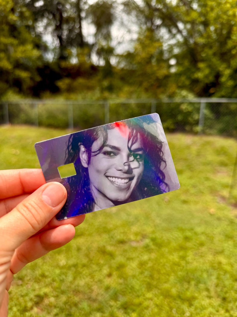 Peut inclure: Une carte rectangulaire en plastique pr&eacute;sentant un portrait photographique en noir et blanc de Michael Jackson. La carte poss&egrave;de une finition holographique qui refl&egrave;te la lumi&egrave;re, cr&eacute;ant des reflets arc-en-ciel sur la surface, et inclut une petite d&eacute;coupe rectangulaire.