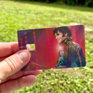 Puede incluir: Una tarjeta rectangular con un retrato sonriente de Elvis Presley con una chaqueta de cuero negra, sobre un fondo rojo. La tarjeta tiene un recorte cerca de la parte superior. La imagen se sostiene contra un fondo de hierba verde.