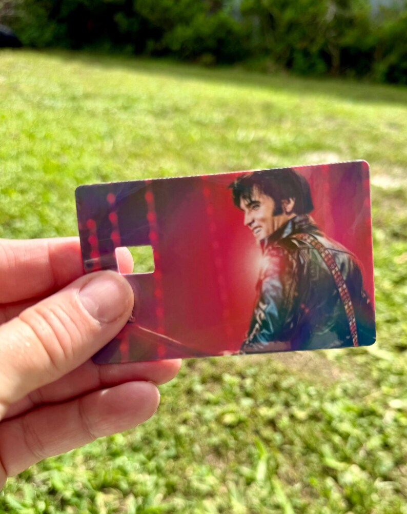 Puede incluir: Una tarjeta rectangular con un retrato sonriente de Elvis Presley con una chaqueta de cuero negra, sobre un fondo rojo. La tarjeta tiene un recorte cerca de la parte superior. La imagen se sostiene contra un fondo de hierba verde.