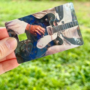 Puede incluir: Una tarjeta rectangular con una guitarra el&eacute;ctrica en blanco y negro siendo tocada por una mano. La tarjeta tiene un fondo azul y blanco y un cuadrado verde en el lado izquierdo. La tarjeta se sostiene contra un fondo verde borroso.