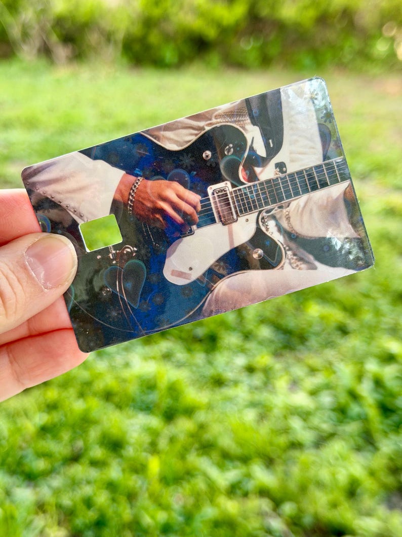 Puede incluir: Una tarjeta rectangular con una guitarra el&eacute;ctrica en blanco y negro siendo tocada por una mano. La tarjeta tiene un fondo azul y blanco y un cuadrado verde en el lado izquierdo. La tarjeta se sostiene contra un fondo verde borroso.