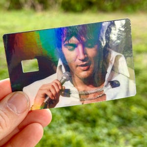 Puede incluir: Una tarjeta hologr&aacute;fica con un retrato de Elvis Presley cantando en un micr&oacute;fono. La imagen muestra a Elvis con una camisa blanca y m&uacute;ltiples collares plateados. La tarjeta tiene forma rectangular con un peque&ntilde;o recorte cuadrado.
