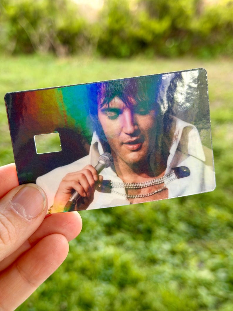 Puede incluir: Una tarjeta hologr&aacute;fica con un retrato de Elvis Presley cantando en un micr&oacute;fono. La imagen muestra a Elvis con una camisa blanca y m&uacute;ltiples collares plateados. La tarjeta tiene forma rectangular con un peque&ntilde;o recorte cuadrado.