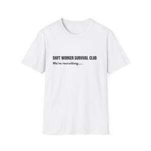 Puede incluir: Camiseta blanca con el texto "SHIFT WORKER SURVIVAL CLUB" y "We're recruiting...." impreso en negro. La camiseta tiene cuello redondo y mangas cortas.