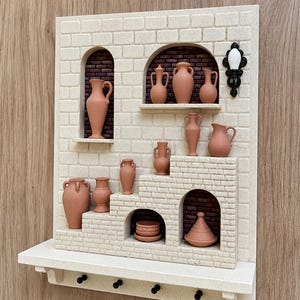 May include: A wall-mounted display featuring a cream-colored brick facade with arched niches and shelves holding miniature terracotta-colored pottery vessels. The structure includes a bottom shelf with four black hooks and a black wall lamp.