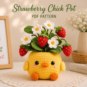 May include: A yellow crocheted chick-shaped pot containing a crocheted strawberry plant with red berries, green leaves, and white flowers. The text Strawberry Chick Pot PDF PATTERN is visible at the top. The chick has black eyes and orange feet.