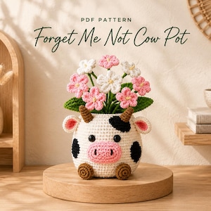 May include: A crochet cow-shaped pot featuring a white body with black spots, a pink snout, brown horns, and small brown feet. The pot contains a bouquet of crocheted pink and white flowers with green leaves. Text reads: PDF PATTERN Forget Me Not Cow Pot.
