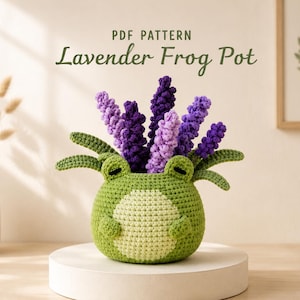 Può includere: Un vaso all'uncinetto a forma di rana verde con una pancia verde chiaro, con rametti di lavanda viola che emergono dalla parte superiore. Il testo PDF PATTERN Lavender Frog Pot è visualizzato sopra l'oggetto in un carattere verde scuro.