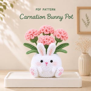 Może przedstawiać: Wzór szydełkowy w formacie PDF o nazwie Carnation Bunny Pot przedstawiający białą doniczkę w kształcie króliczka z różowymi łapkami i rumieńcami, zawierającą cztery różowe goździki z zielonymi łodygami i liśćmi z włóczki.