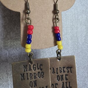 May include: A pair of gold-toned earrings with a square charm that reads "Magic Mirror on the Wall" and "Fairest One of All". The charms are attached to a chain with red, blue, and yellow beads.