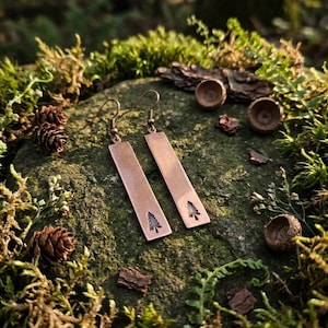 May include: Copper-colored rectangular earrings with a tree design. The earrings are displayed on a mossy rock with pinecones and acorns in the background. The earrings have a simple, nature-inspired design.