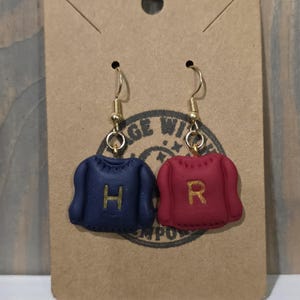 Puede incluir: Pendientes hechos a mano con colgantes de suéter en miniatura. Uno es azul marino con una "H" dorada, el otro rojo con una "R" dorada. Los pendientes cuelgan de ganchos dorados.
