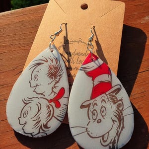 May include: A pair of teardrop-shaped earrings featuring illustrations of characters from a children's book. One earring depicts a character with a red and white striped hat, while the other shows two other characters. The earrings are attached to silver-colored hooks.