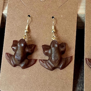 May include: Three pairs of frog-shaped earrings displayed on brown cardstock. The earrings are brown with varying metal hook colors: silver, gold, and dark bronze. The frog design is detailed, with visible legs and a textured surface.