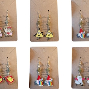 May include: Six pairs of dangle earrings on brown cards. The earrings feature cartoon characters in various designs, including gnomes, books, and geometric shapes. The earrings have gold-colored hooks and decorative charms.