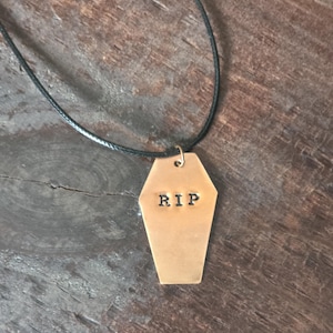 Puede incluir: Un collar con colgante en forma de ataúd de color cobre con las letras "RIP" estampadas. El colgante cuelga de un cordón negro. El fondo es una superficie de madera oscura.