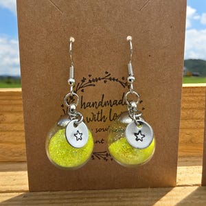 May include: A pair of handmade earrings featuring clear glass globes filled with yellow glitter. Each globe hangs from a silver-colored hook and has a small, round charm with a star cutout. The earrings are displayed on a brown card.