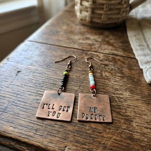 May include: Copper earrings with square pendants, each stamped with text. One reads "I'LL GET YOU," the other "MY PRETTY." The earrings have beaded accents in black, green, and yellow, and a mix of colorful beads. The earrings are displayed on a wooden surface.