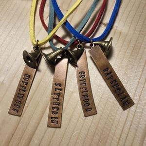 May include: Four necklaces with different colored cords and charms. Each charm is a small bronze-colored hat with a house name from the Harry Potter series: Gryffindor, Slytherin, Hufflepuff, and Ravenclaw.