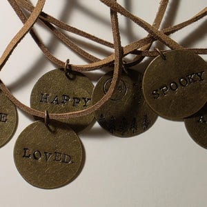 May include: Six brown leather necklaces with round metal charms. The charms have the words "Nope", "Happy", "Loved", "Spooky", "Away", and a moon and trees design.