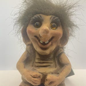 May include: A whimsical troll figurine with a smiling expression. The troll has a large nose, prominent ears, and a wild, gray-brown hair. It is wearing a simple, textured outfit and has a light brown skin tone.