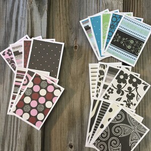 Puede incluir: Una colección de tarjetas de felicitación estampadas con varios diseños, incluyendo lunares, patrones florales y formas geométricas. Las tarjetas tienen un borde blanco y están dispuestas sobre una superficie de madera envejecida.