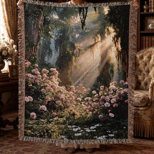 May include: A woven throw blanket featuring a detailed landscape scene of a lush forest with blooming pink flowers and white daisies in the foreground. Sunlight streams through the dense canopy of trees, creating bright rays of light.