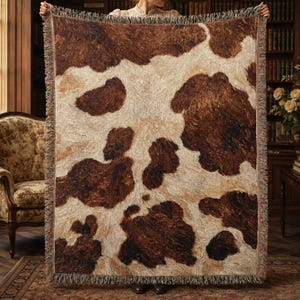 May include: A rectangular woven throw blanket featuring a brown and cream cowhide pattern. The textile displays irregular dark brown patches against a lighter cream-colored background with a textured, woven surface and fringed edges on all sides.