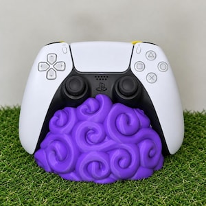 Può includere: Un controller per videogiochi bianco e nero appoggiato su un supporto viola. Il supporto presenta un design testurizzato con motivi a vortice simili a nuvole. Il controller ha un logo PlayStation centrale, due levette analogiche nere e un D-pad.