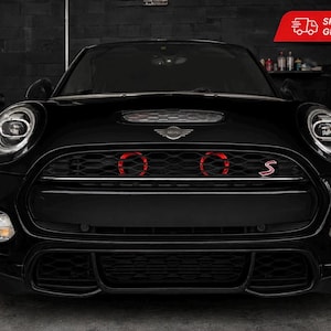 Könnte beinhalten: Ein schwarzer Mini Cooper S von vorne gesehen, mit einem schwarzen Wabengrill, zwei kreisförmigen roten Akzentlichtern, einem silbernen Mini-Emblem, einem roten S-Emblem, runden LED-Scheinwerfern und Nebelscheinwerfern in einer dunklen Garage.