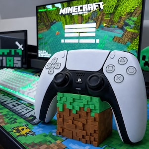 Pode incluir: Um controle de jogo sem fio branco e preto repousa sobre um suporte em forma de bloco marrom e verde. O suporte apresenta uma textura pixelizada de grama e terra. Um monitor de computador exibe o menu do jogo Minecraft Java Edition.