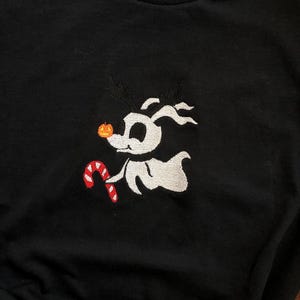 Puede incluir: Camiseta negra con un dise&ntilde;o bordado de un perro de dibujos animados. El perro tiene cuernos, una nariz de calabaza y un bast&oacute;n de caramelo. El perro es blanco con cuernos negros y un bast&oacute;n de caramelo rojo y blanco. La camiseta es de algod&oacute;n pesado.