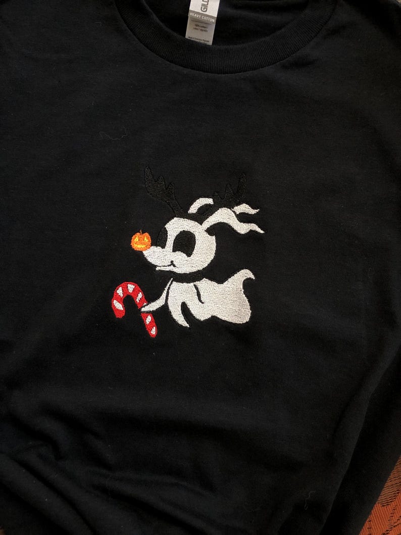 Puede incluir: Camiseta negra con un dise&ntilde;o bordado de un perro de dibujos animados. El perro tiene cuernos, una nariz de calabaza y un bast&oacute;n de caramelo. El perro es blanco con cuernos negros y un bast&oacute;n de caramelo rojo y blanco. La camiseta es de algod&oacute;n pesado.
