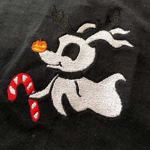 Puede incluir: Dise&ntilde;o bordado de un perro de dibujos animados con cuernos, una nariz de calabaza y un bast&oacute;n de caramelo. El perro es blanco con detalles negros sobre un fondo negro. El bast&oacute;n de caramelo es rojo y blanco.