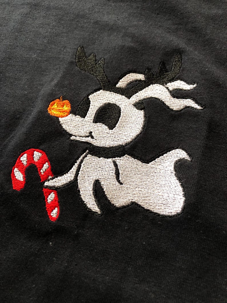 Puede incluir: Dise&ntilde;o bordado de un perro de dibujos animados con cuernos, una nariz de calabaza y un bast&oacute;n de caramelo. El perro es blanco con detalles negros sobre un fondo negro. El bast&oacute;n de caramelo es rojo y blanco.