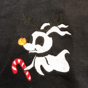 Puede incluir: Tela negra con un dise&ntilde;o bordado de un personaje blanco fantasmal con cuernos negros, una nariz de calabaza y un bast&oacute;n de caramelo rojo y blanco. El personaje es de la pel&iacute;cula "Pesadilla antes de Navidad".