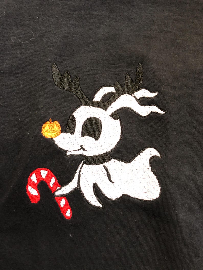 Puede incluir: Tela negra con un dise&ntilde;o bordado de un personaje blanco fantasmal con cuernos negros, una nariz de calabaza y un bast&oacute;n de caramelo rojo y blanco. El personaje es de la pel&iacute;cula "Pesadilla antes de Navidad".