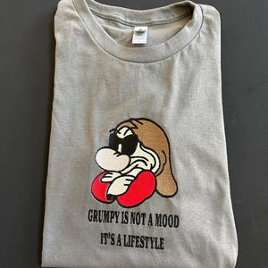 Peut inclure: T-shirt gris avec un dessin de personnage de dessin animé. Le personnage a des cheveux bruns, des lunettes de soleil et des gants rouges. Le texte "GRUMPY IS NOT A MOOD IT'S A LIFESTYLE" est imprimé en dessous.