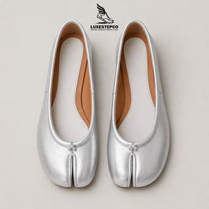 May include: A pair of silver ballet flats with a split-toe design. The shoes have a smooth, metallic finish and a small bow detail at the toe. The interior is white with a tan insole.
