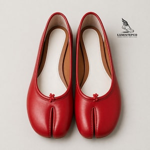 Op de afbeelding: Een paar rode leren ballerina's met een split-teenontwerp. De schoenen hebben een witte binnenzool en een klein knoopdetail aan de bovenkant van de split. Het merklogo "Luxestepco" is zichtbaar.