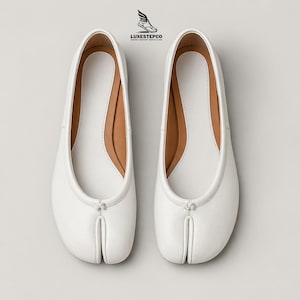 May include: A pair of white leather ballet flats with a split-toe design. The shoes have a minimalist aesthetic with a light brown insole and a small bow detail at the top. The logo "LUXESTEPCO" is visible at the top.